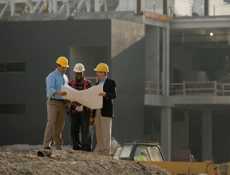 Three construction workers in hard hats reviewing blueprints at a construction site with heavy machinery in the background.