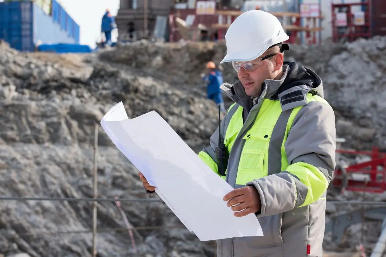 A construction worker in a hard hat and reflective vest examines architectural plans at a construction site.