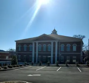 A two-story brick building with large white columns and a central cupola, situated under a clear blue sky with sunlight streaming down.