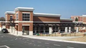 A modern brick building with large windows and a connecting covered walkway, situated next to a parking area under a clear sky.